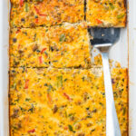 A baked sausage, egg, and cheese casserole with vegetables and sausage, sliced into squares in a white baking dish. A metal spatula rests under one of the slices.