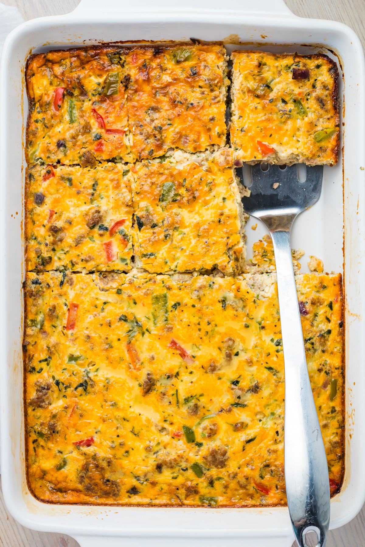 A baked sausage, egg, and cheese casserole with vegetables and sausage, sliced into squares in a white baking dish. A metal spatula rests under one of the slices.