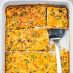 A baked sausage, egg, and cheese casserole, cut into squares in a white baking dish, with one piece being lifted by a metal spatula.