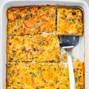 A baked sausage, egg, and cheese casserole, cut into squares in a white baking dish, with one piece being lifted by a metal spatula.