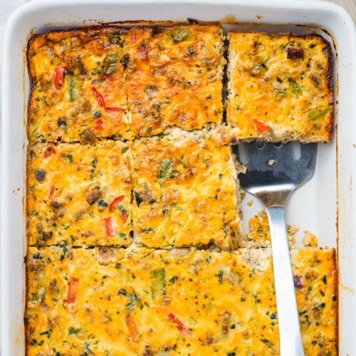 A baked sausage, egg, and cheese casserole, cut into squares in a white baking dish, with one piece being lifted by a metal spatula.