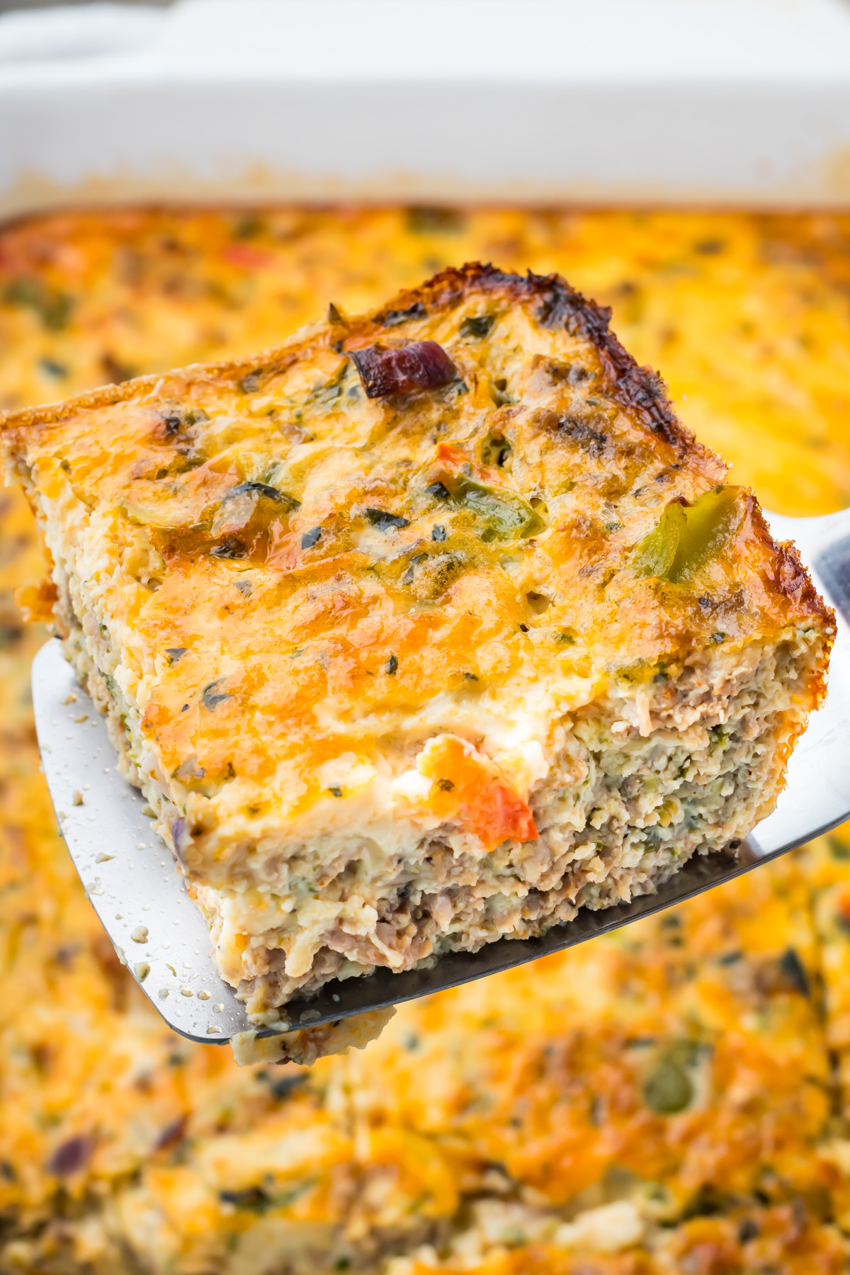 A metal spatula holding a square slice of baked sausage, egg, and cheese casserole with vegetables, above the remaining casserole in a baking dish.