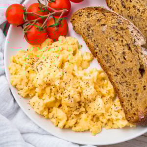 A plate with scrambled eggs (without milk) topped with black pepper, two slices of whole grain toast, and a cluster of vine tomatoes.