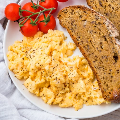 A plate with scrambled eggs (without milk) topped with black pepper, two slices of whole grain toast, and a cluster of vine tomatoes.