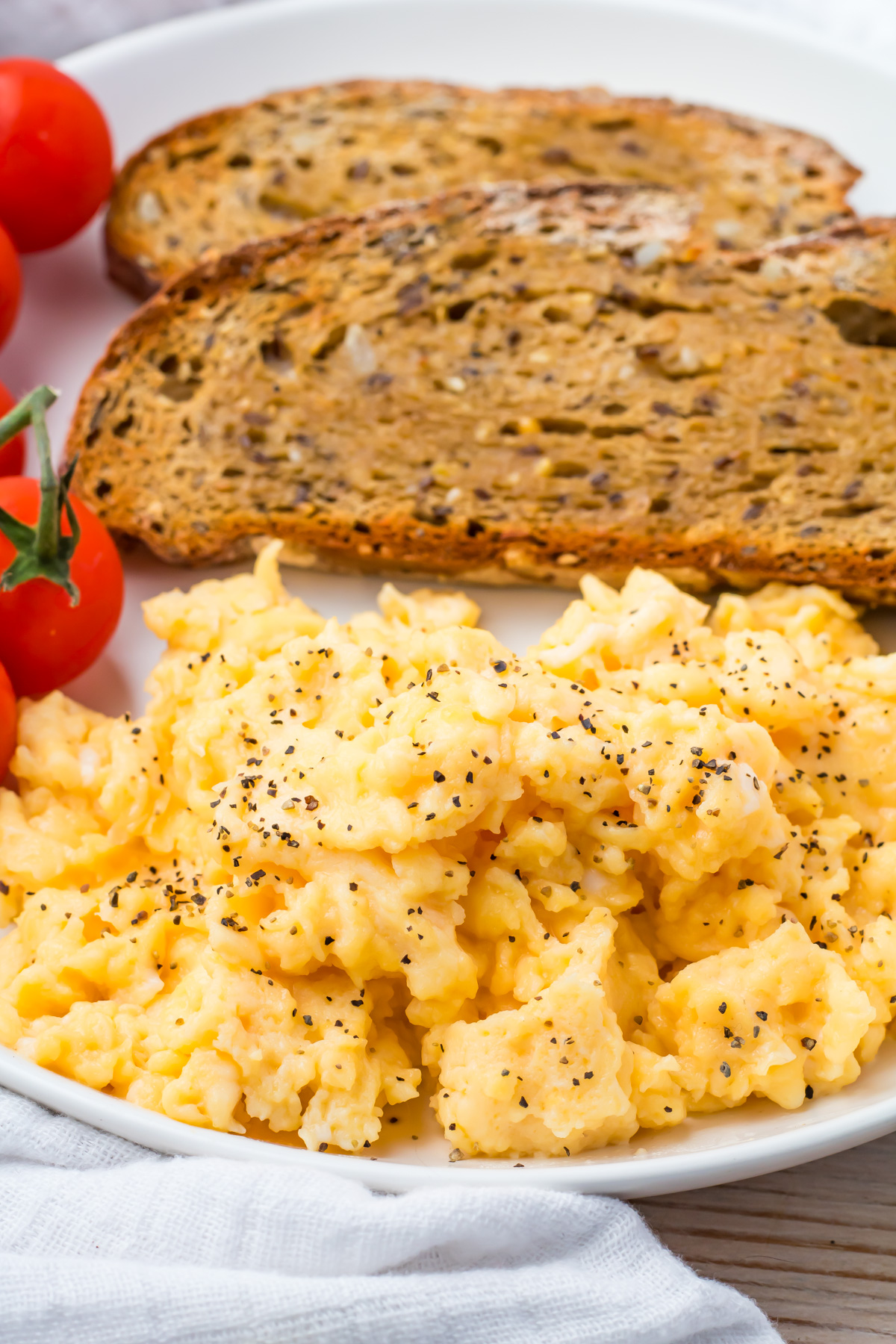 A plate with scrambled eggs (without milk) sprinkled with black pepper, two slices of toasted multigrain bread, and a small bunch of cherry tomatoes.