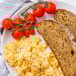 A plate with scrambled eggs (without milk), two slices of whole grain toast, and a small bunch of vine tomatoes, set on a white cloth.