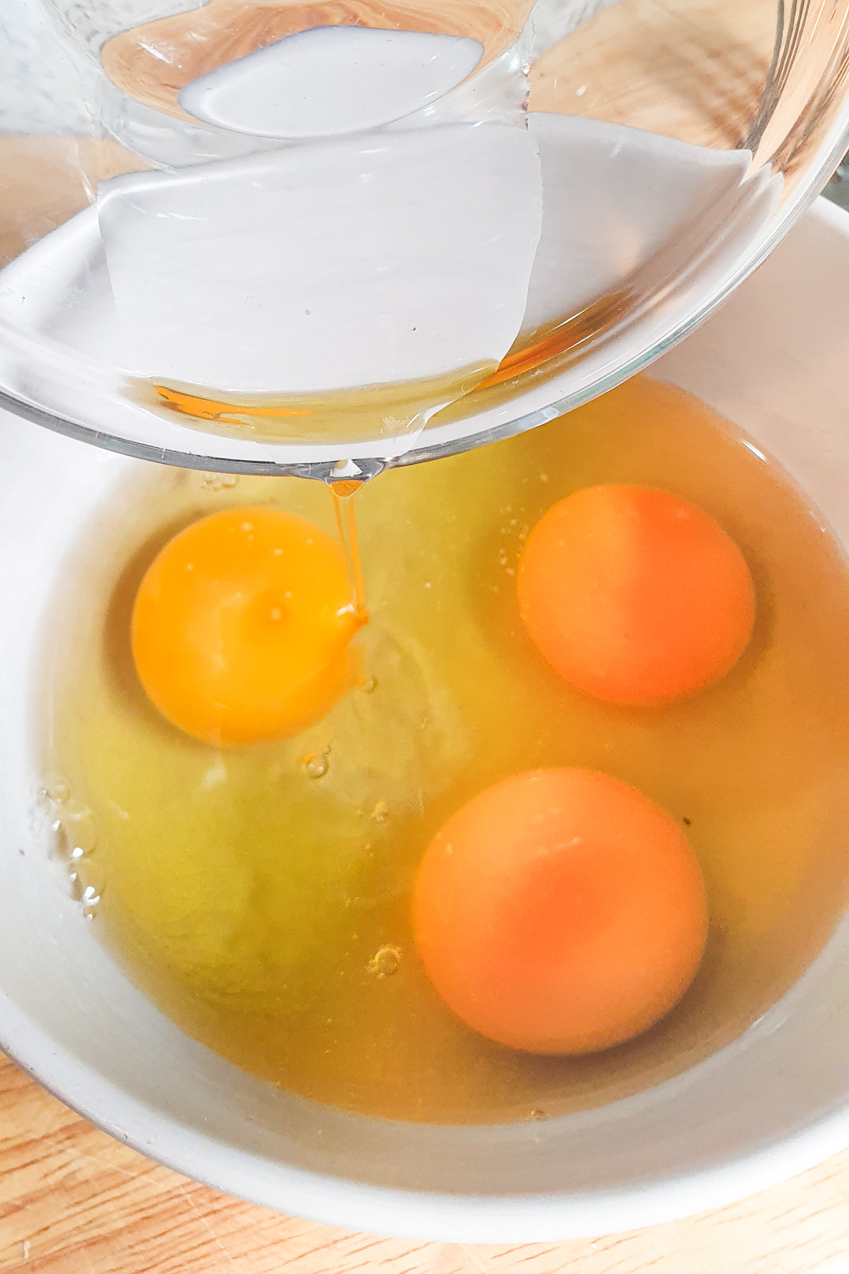 A clear bowl is used to pour water into a white bowl that already contains three cracked eggs.