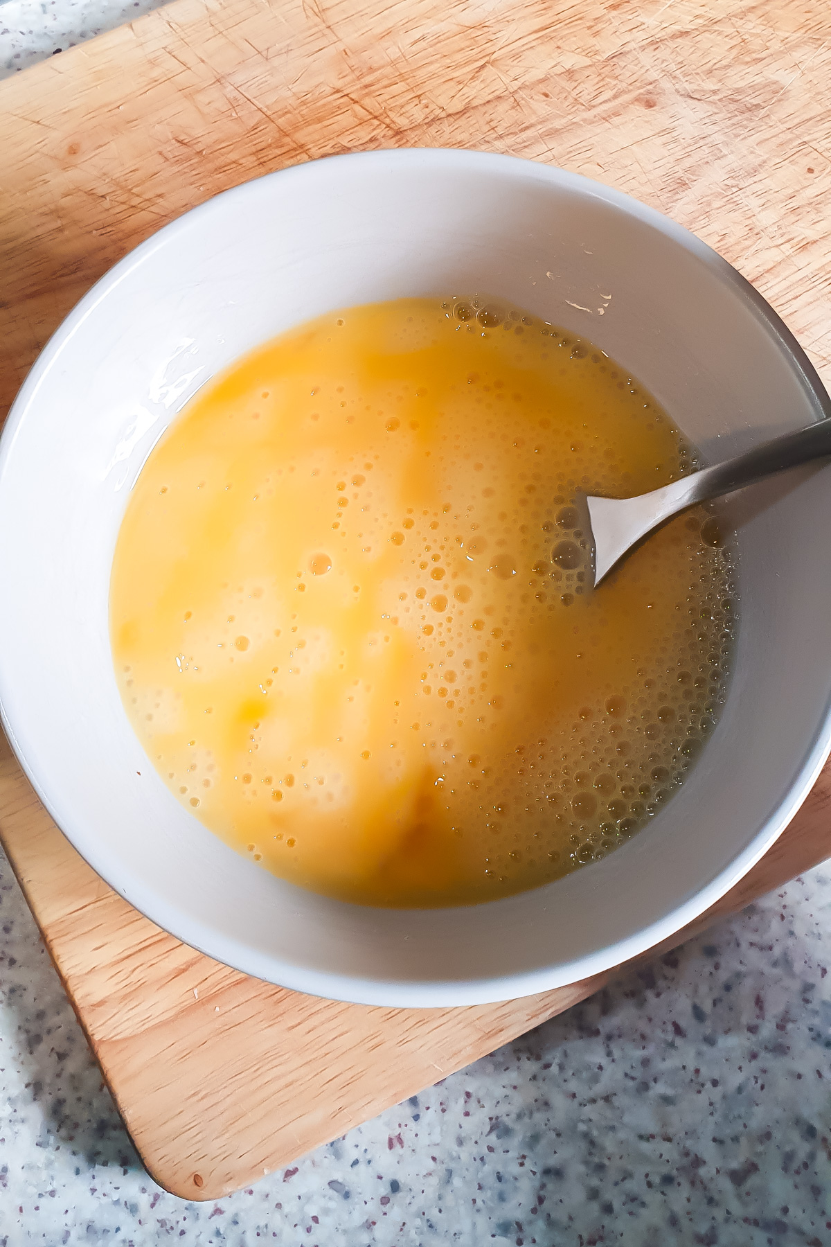 A white bowl containing beaten eggs with a metal spoon, placed on a wooden cutting board on a light-colored countertop.