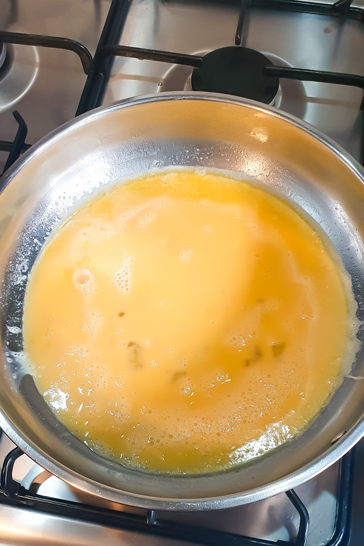 A stainless steel pan with beaten eggs cooking on a gas stove, beginning to set around the edges.