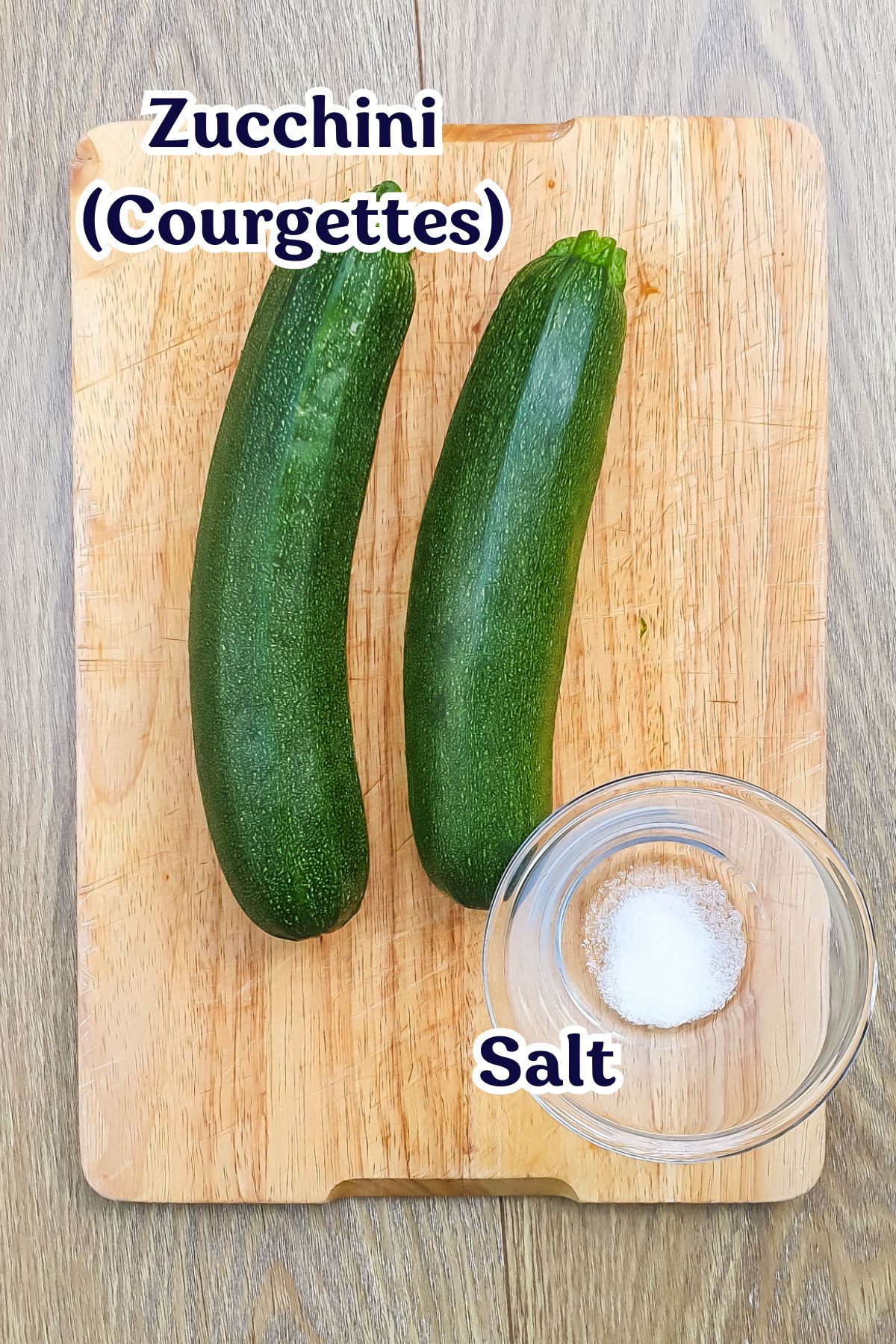 Two zucchinis on a wooden cutting board next to a small glass bowl filled with salt.