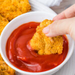A hand dips a breaded air fryer ground chicken nugget into a bowl of ketchup, with more chicken nuggets on a white plate nearby.