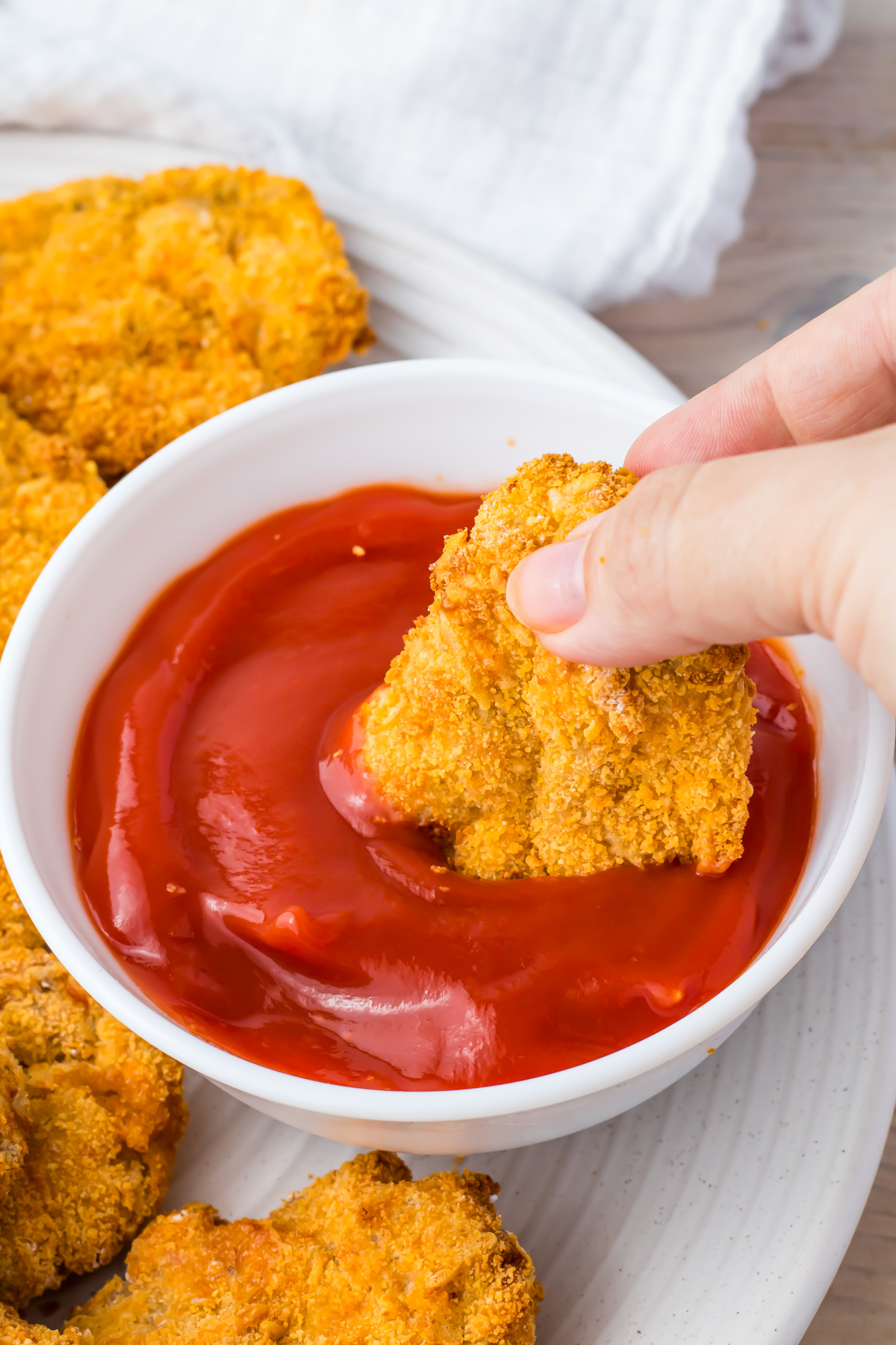 A hand dips a breaded air fryer ground chicken nugget into a bowl of ketchup, with more chicken nuggets on a white plate nearby.