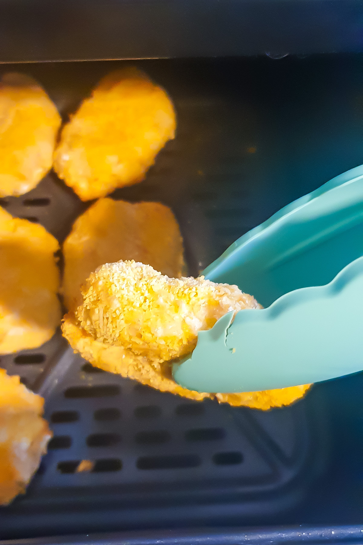A pair of teal tongs holds a breaded ground chicken nugget above several more nuggets inside an air fryer basket.