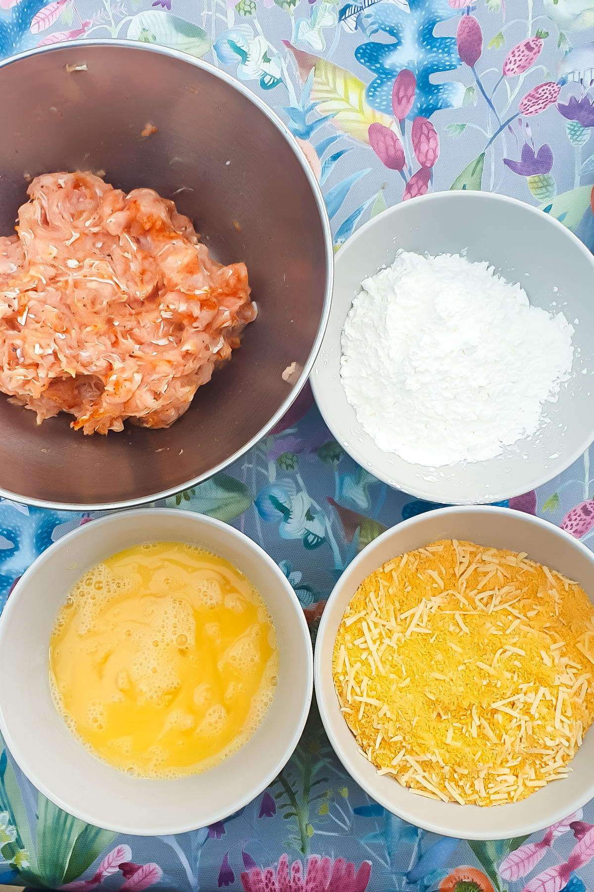 Four bowls on a patterned table: one with ground chicken, one with flour, one with beaten eggs, and one with breadcrumbs and shredded cheese.