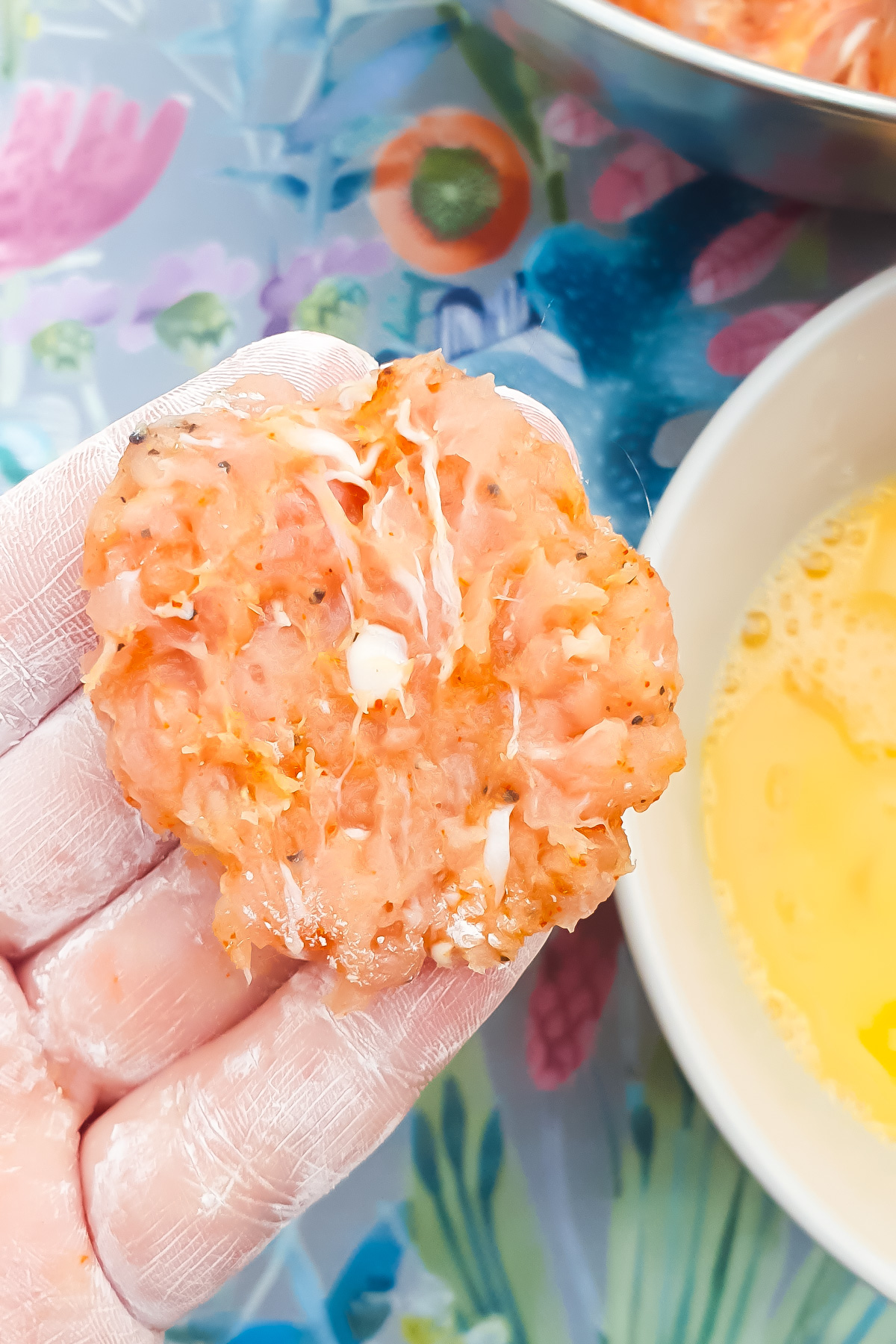 A hand holds a raw, seasoned chicken patty over a bowl with beaten eggs, with a floral-patterned surface underneath.