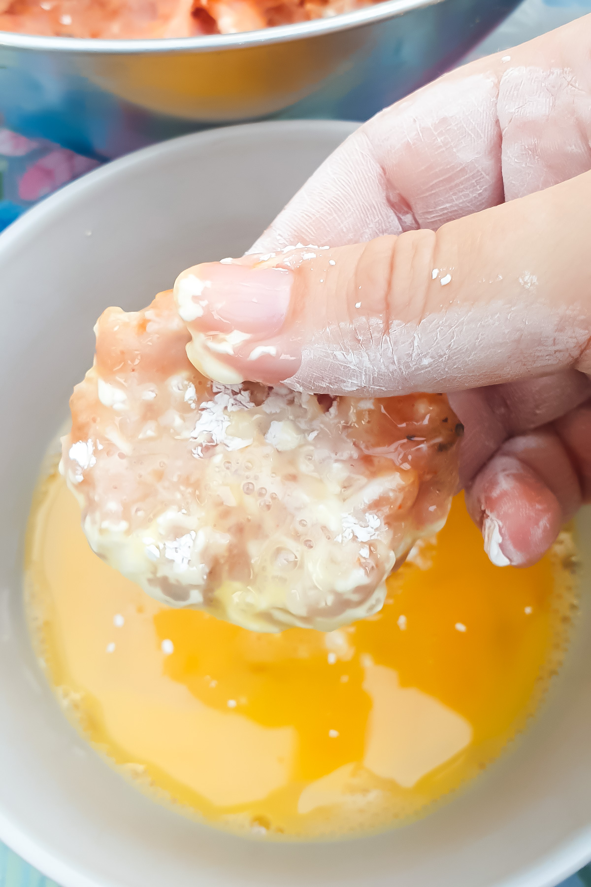 A hand dips a floured piece of ground chicken into a bowl of beaten eggs, preparing it for breading.