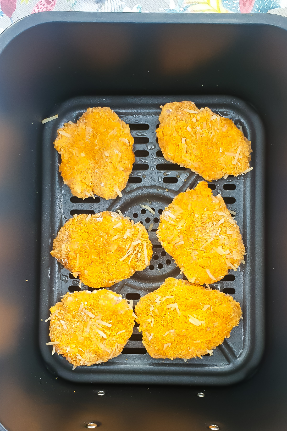 Six breaded ground chicken nuggets are arranged in a single layer inside an air fryer basket, ready to be cooked.