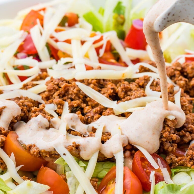 Close-up of a taco salad with ground meat, shredded cheese, sliced cherry tomatoes, lettuce, and creamy Mexican Salad dressing being poured on top.