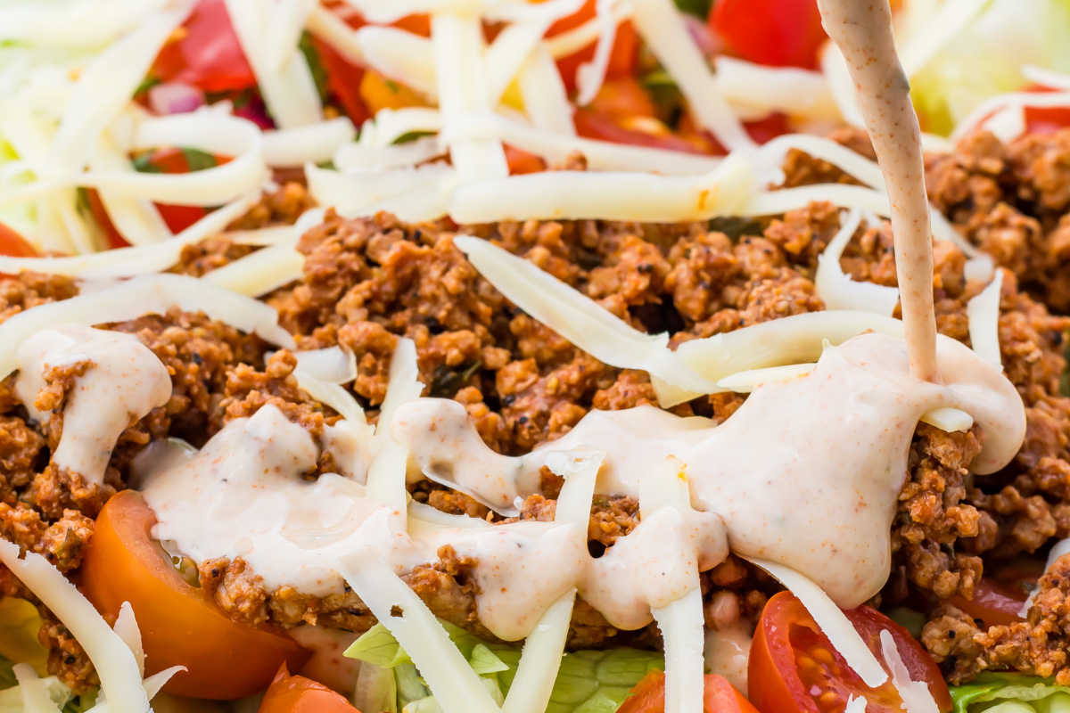 Close-up of a taco salad topped with ground beef, shredded cheese, sliced tomatoes, and a creamy dressing being poured over the ingredients.