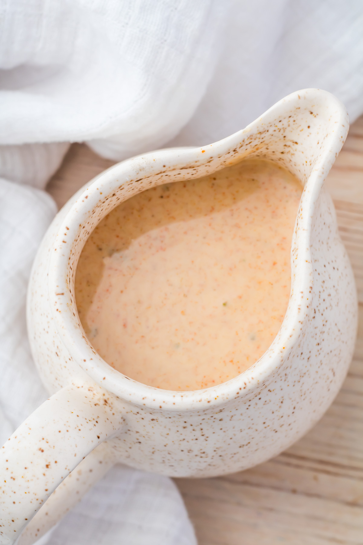 A speckled ceramic pitcher filled with a creamy, Mexican Salad dressing sits on a wooden surface next to a white cloth.