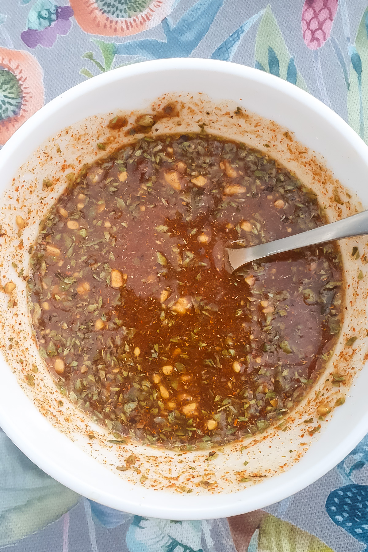 A white bowl containing ingredients and a metal spoon resting inside. The bowl sits on a colorful, patterned surface.