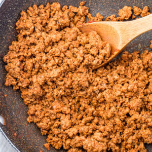 Cooked Mexican taco meat with taco seasoning in a pan, being stirred with a wooden spoon.