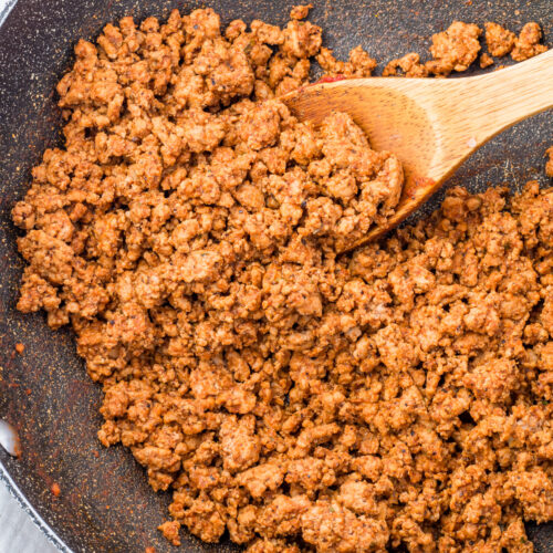 Cooked Mexican taco meat with taco seasoning in a pan, being stirred with a wooden spoon.