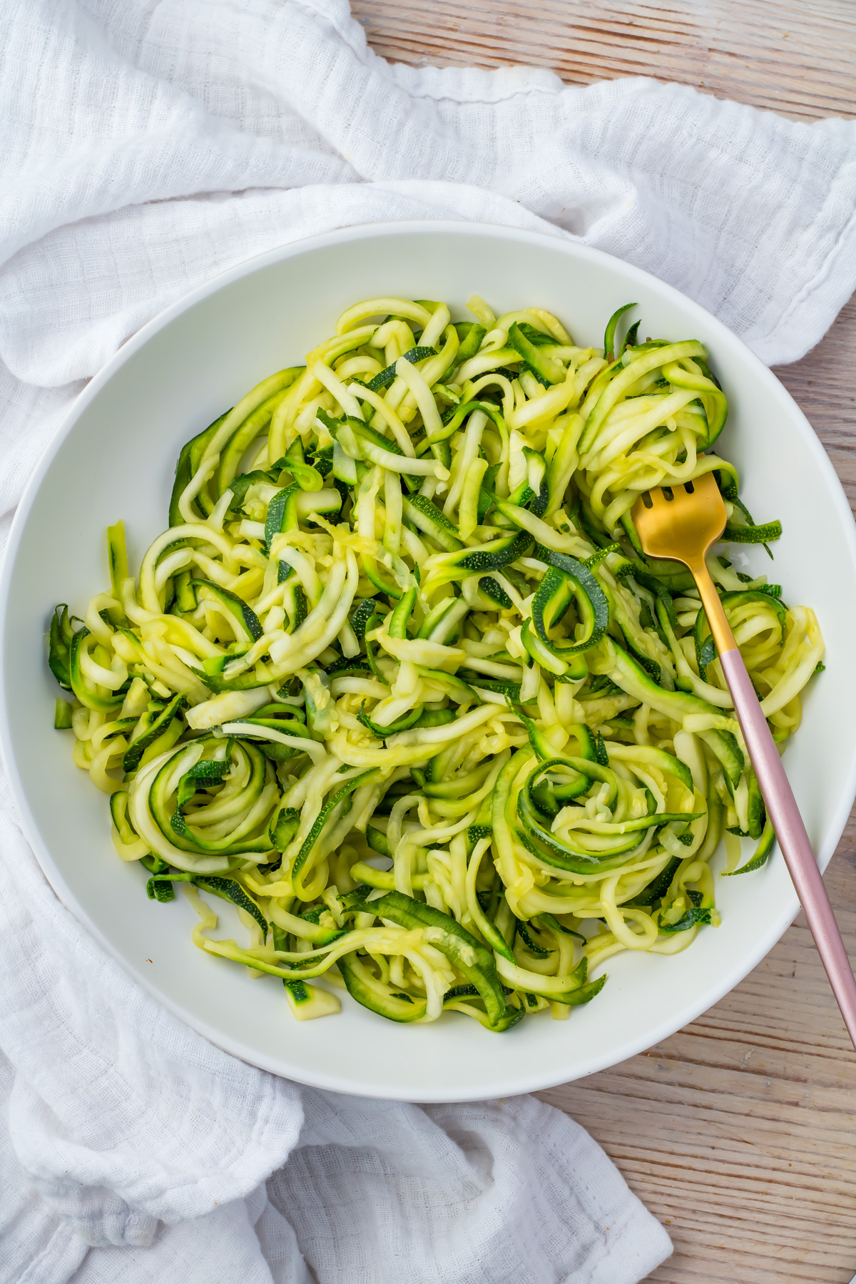 A white bowl filled with spiralized microwave zucchini noodles, next to a gold fork on a white cloth.