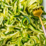 A close-up of spiralized microwave zucchini noodles in a white dish with a gold fork.