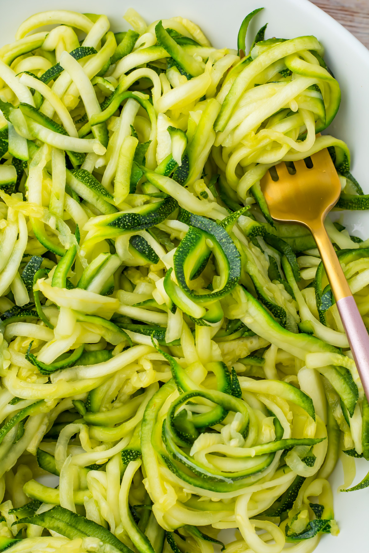 A close-up of spiralized microwave zucchini noodles in a white dish with a gold fork.