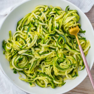 A white bowl filled with spiralized microwave zucchini noodles, with a gold fork resting on the side.