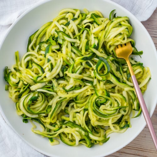 A white bowl filled with spiralized microwave zucchini noodles, with a gold fork resting on the side.