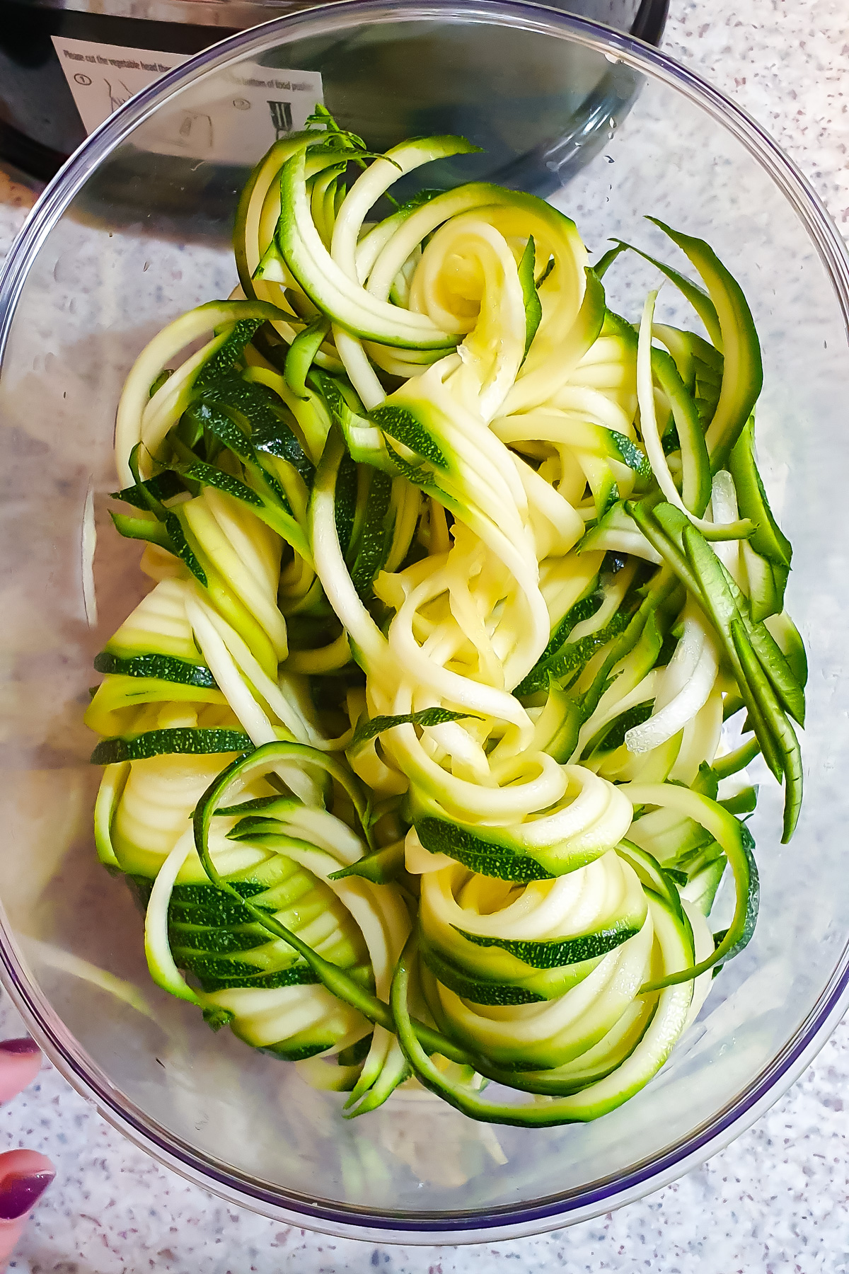 A clear bowl filled with raw zucchini noodles, also known as zoodles, on a speckled countertop.
