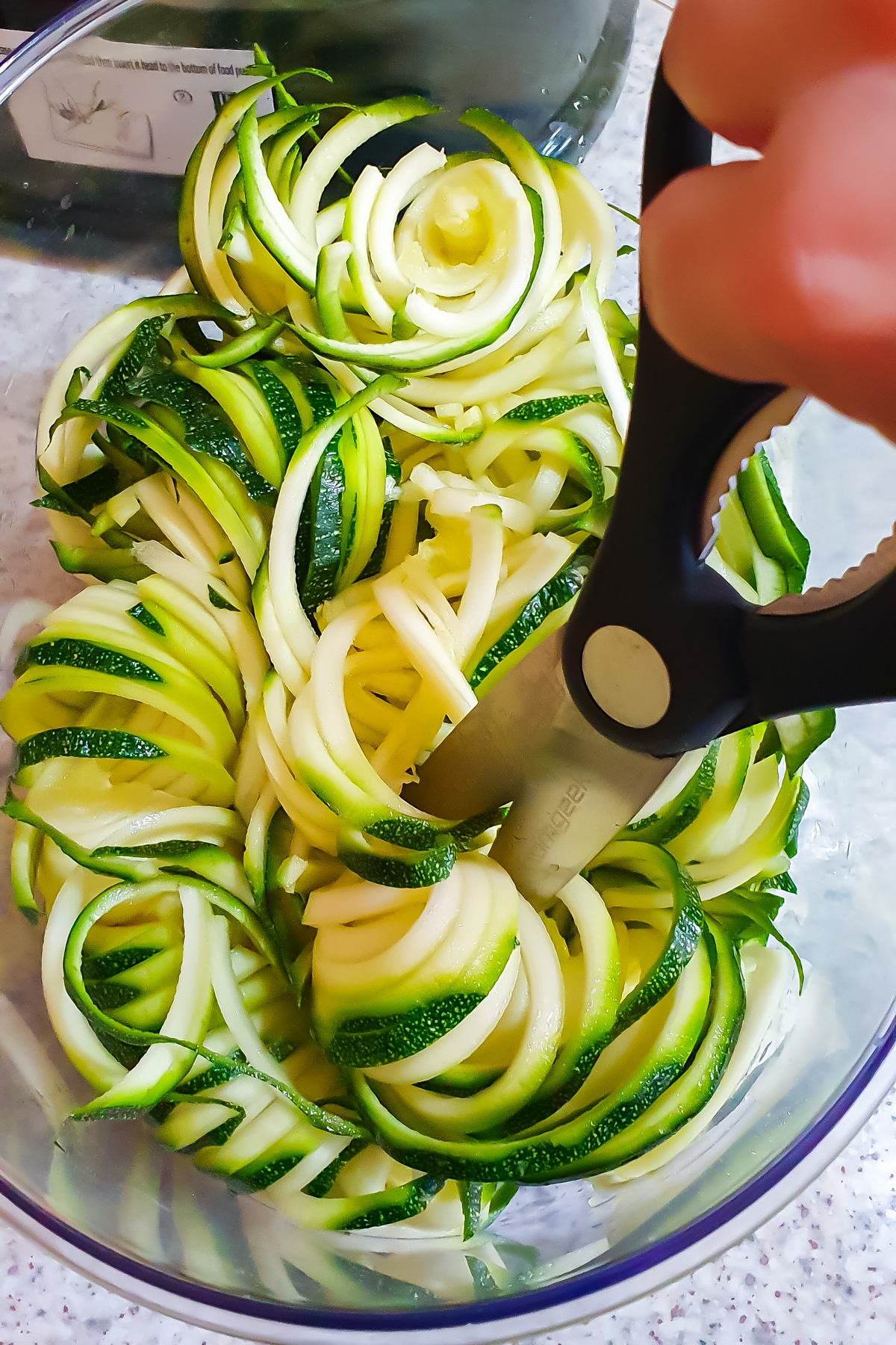 A hand uses kitchen scissors to cut spiralized zucchini noodles in a clear bowl on a countertop.