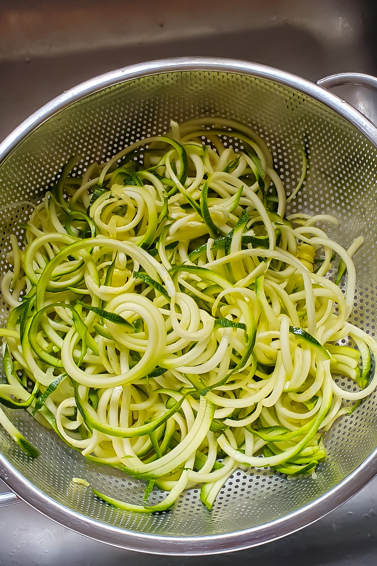 A metal colander filled with freshly spiralized zucchini noodles, known as zoodles, sits in a sink.