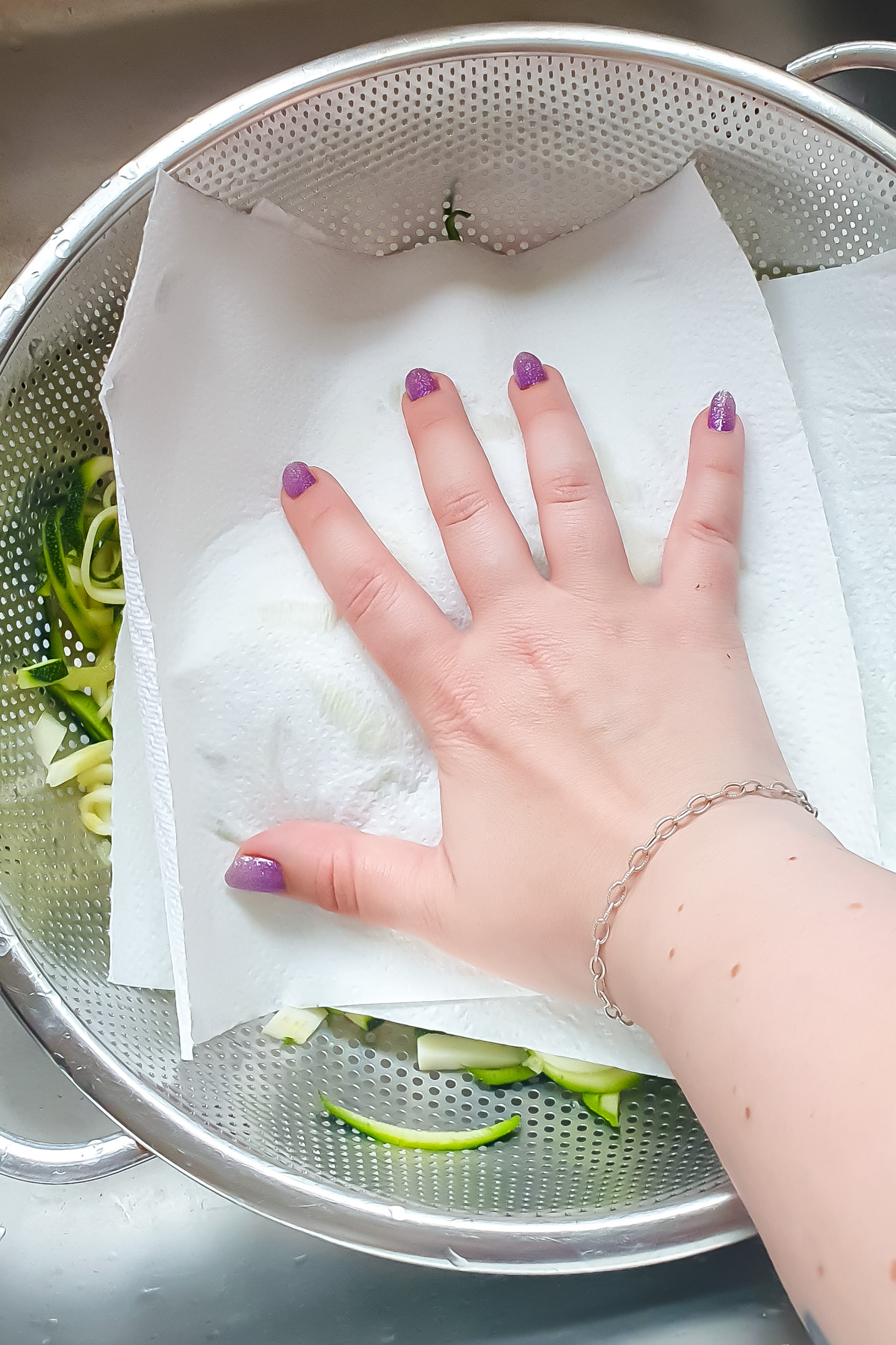 A hand presses paper towels onto spiralized zucchini in a colander, absorbing excess moisture.