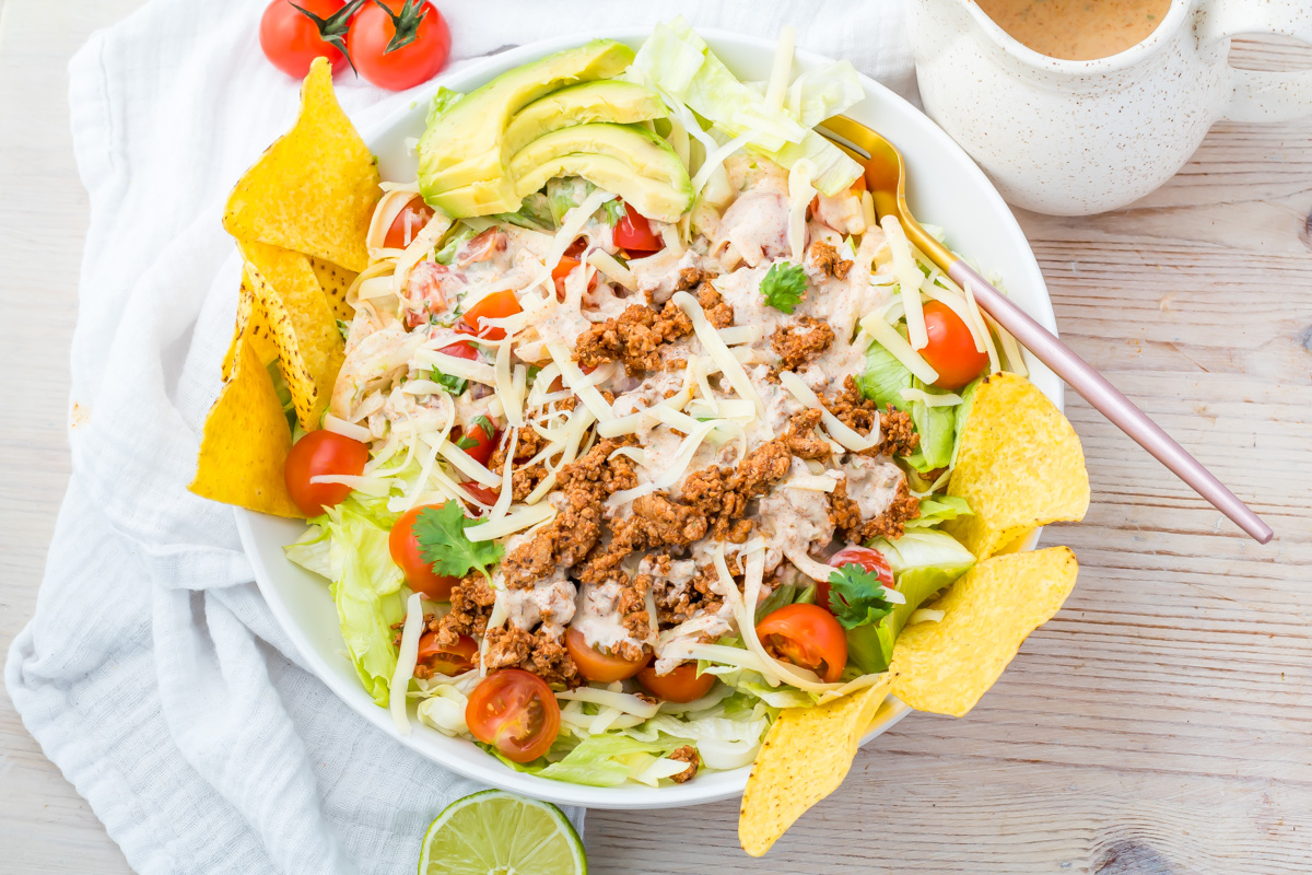 A bowl of taco salad bowl with ground meat, shredded cheese, cherry tomatoes, lettuce, avocado, tortilla chips, and creamy dressing, garnished with cilantro.