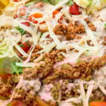 A close-up of a taco salad bowl with ground meat, shredded cheese, cherry tomatoes, avocado slices, lettuce, and creamy dressing.