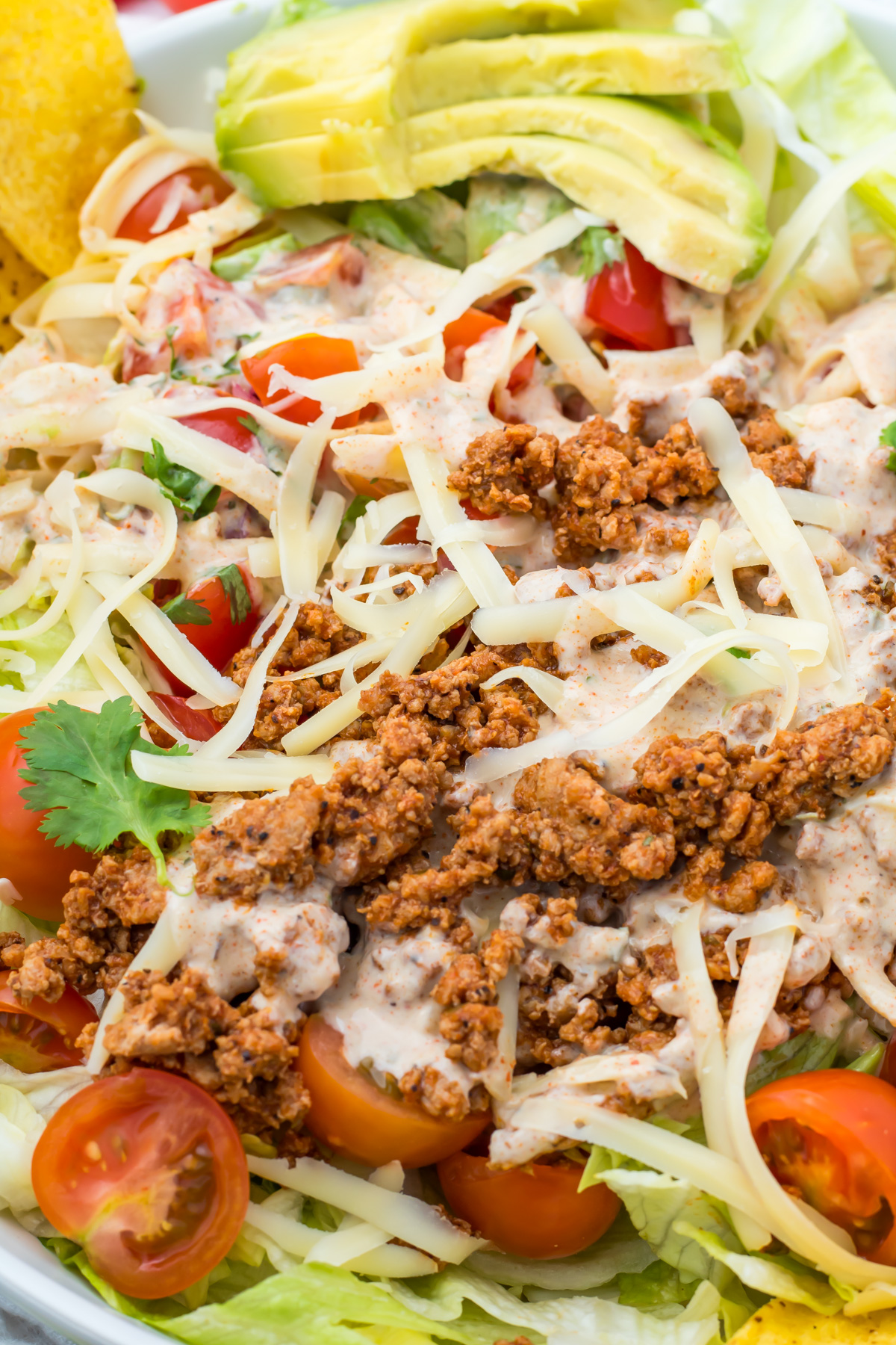 A close-up of a taco salad bowl with ground meat, shredded cheese, cherry tomatoes, avocado slices, lettuce, and creamy dressing.