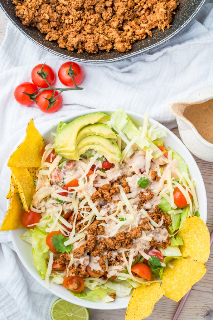 A taco salad with ground meat, shredded cheese, sliced avocado, cherry tomatoes, lettuce, tortilla chips, dressing, and lime on a white plate, with extra meat and dressing nearby.