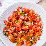 A white bowl filled with chopped taco salsa mixed with red onion and herbs, placed on a white cloth with a halved lime and cherry tomatoes nearby.