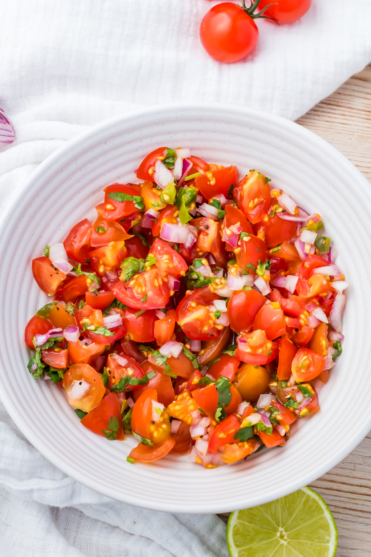 A white bowl filled with chopped taco salsa mixed with red onion and herbs, placed on a white cloth with a halved lime and cherry tomatoes nearby.