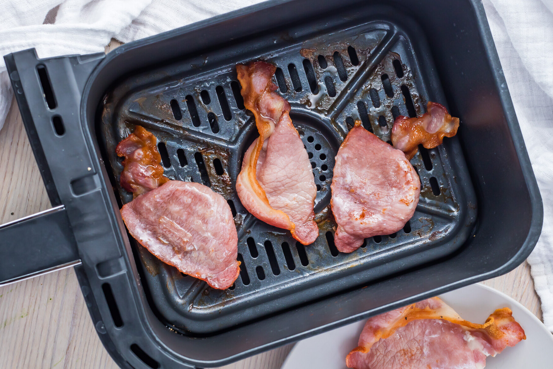 Slices of cooked Canadian bacon nestled inside an air fryer basket, with a piece of savory bacon resting on a white plate beside it atop a light wooden surface.