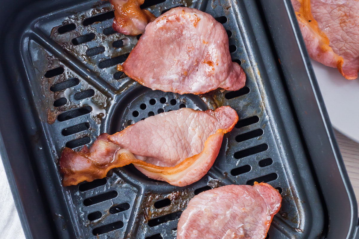 Slices of cooked Canadian bacon nestled inside an air fryer basket, with a piece of savory bacon resting on a white plate beside it atop a light wooden surface.