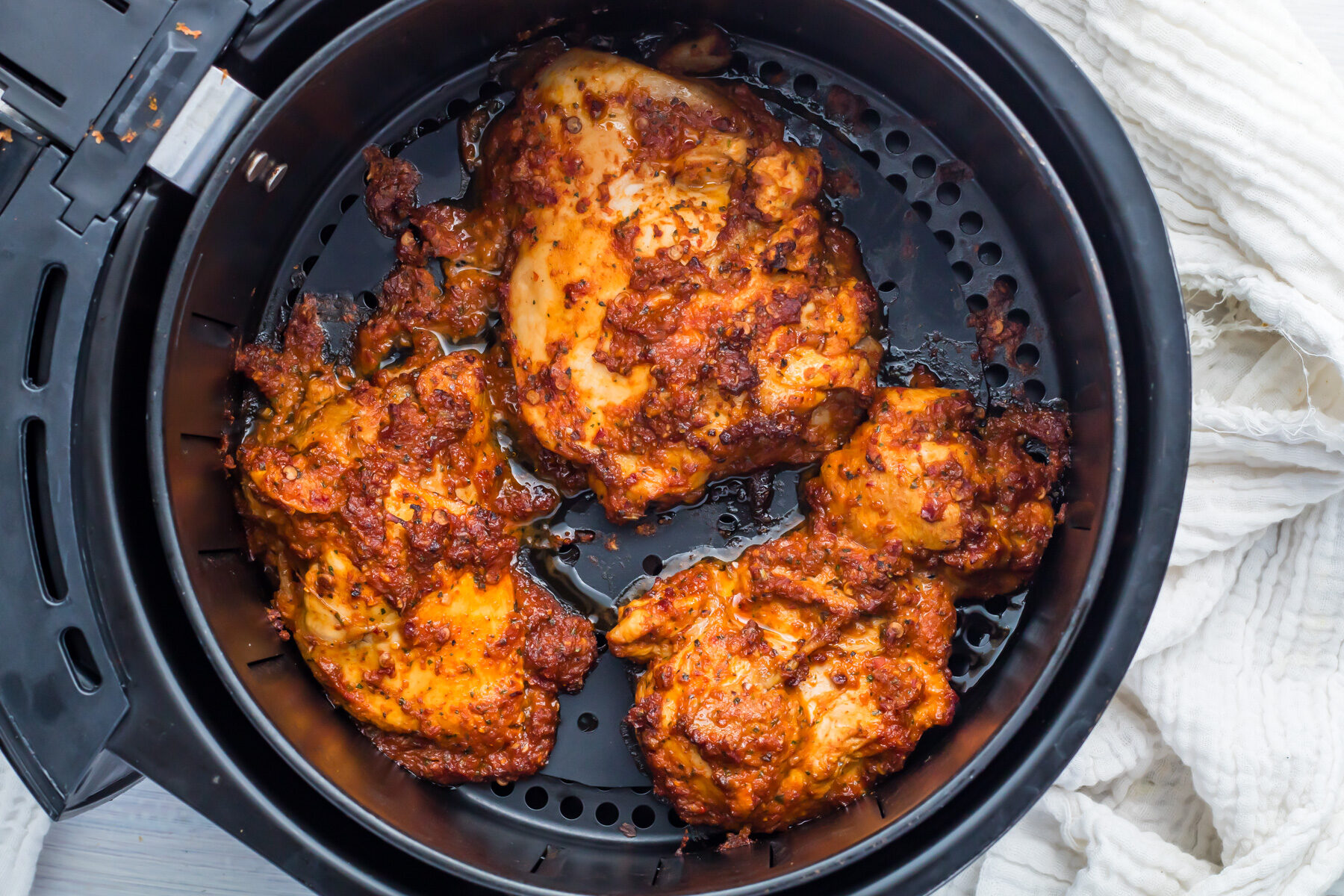 Inside the black air fryer basket, three pieces of pollo asado rest, boasting a rich, golden-brown hue with a visible layer of spices. The succulent chicken stands out against the partially visible light-colored cloth in the background.