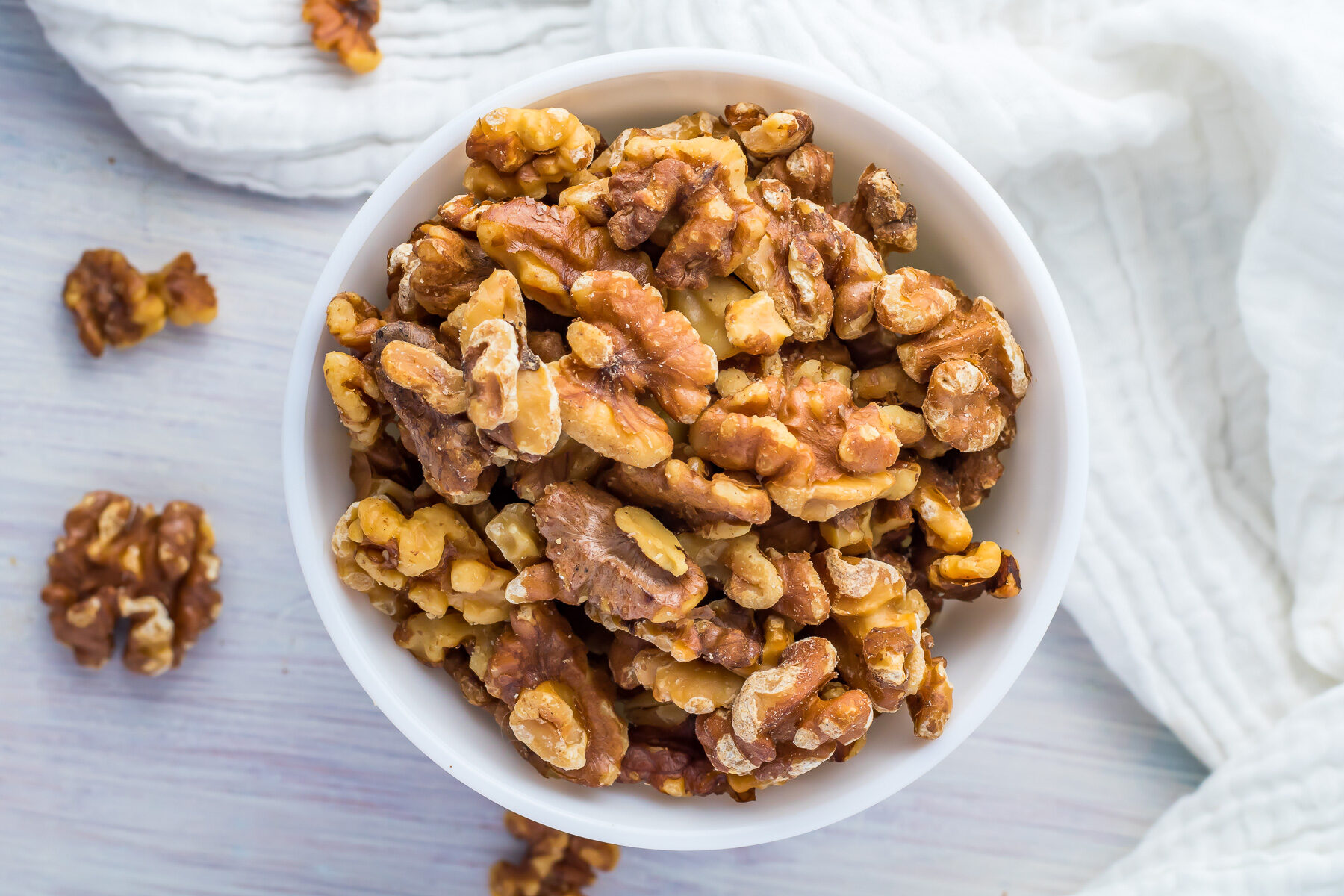 Over head shot of air fryer roasted walnuts in a white bowl with walnuts scattered on the table and a white cloth.