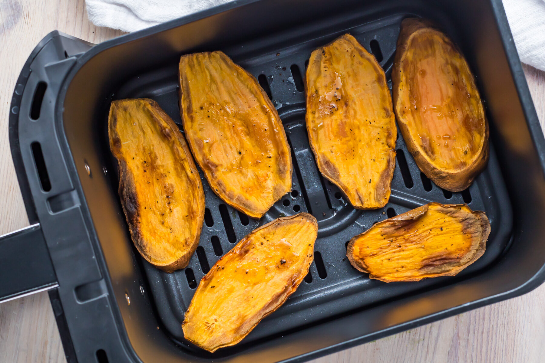 Slices of roasted sweet potato toast in an air fryer basket, with a golden-brown finish. The background shows a light wooden surface and a white cloth peeking from the corner.