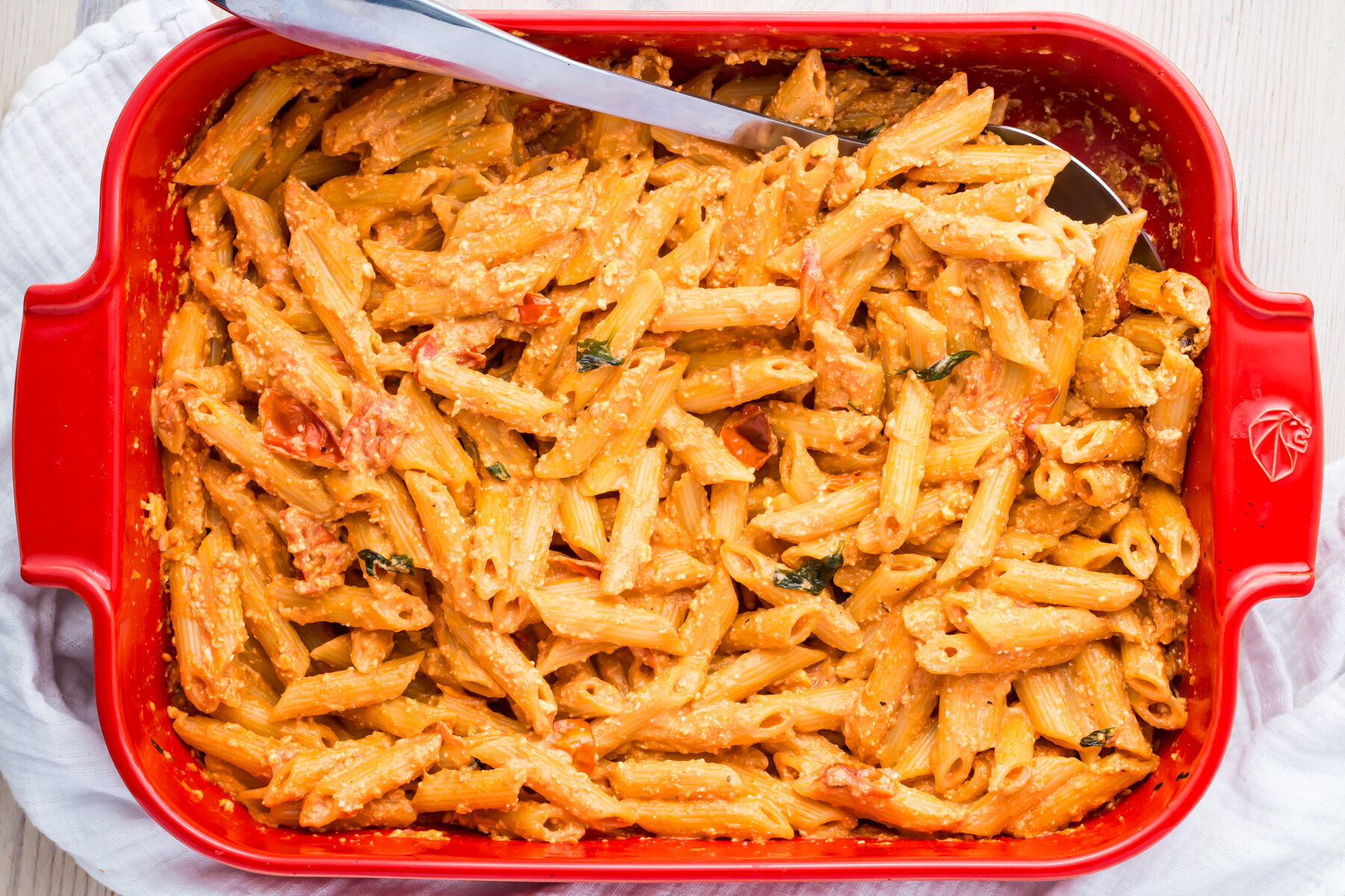 A red baking dish filled with creamy baked penne pasta, mixed with a rich sauce and garnished with herbs. A serving spoon is placed on the left side of the dish.