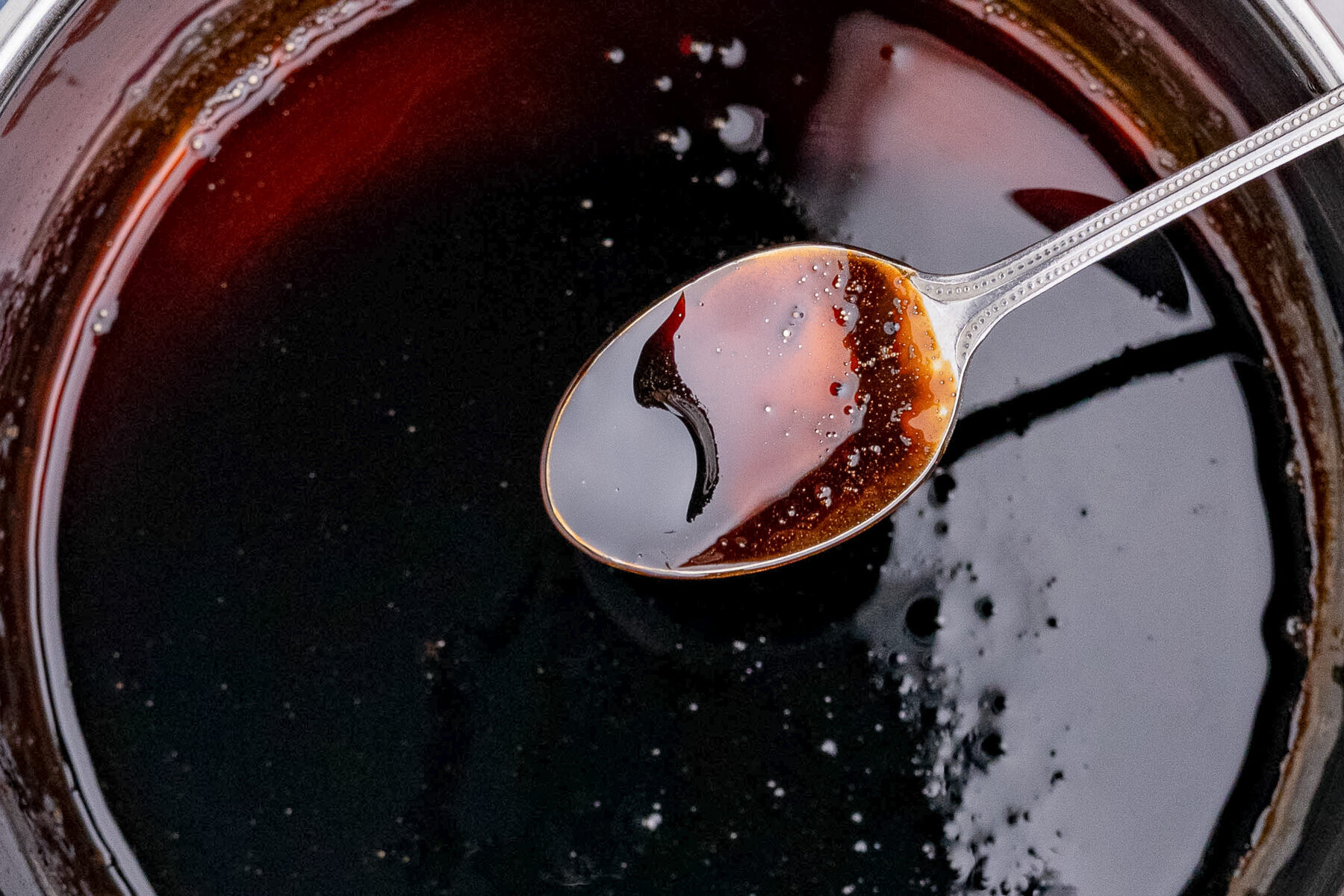 A silver spoon is dipped into a dark, thick balsamic glaze in a pot, reflecting light and showing a glossy surface with small bubbles and a hint of red.