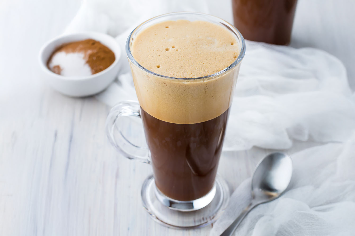 2 Greek frappes in tall glasses on a white table next to a white cloth and a small bowl of coffee and sugar.